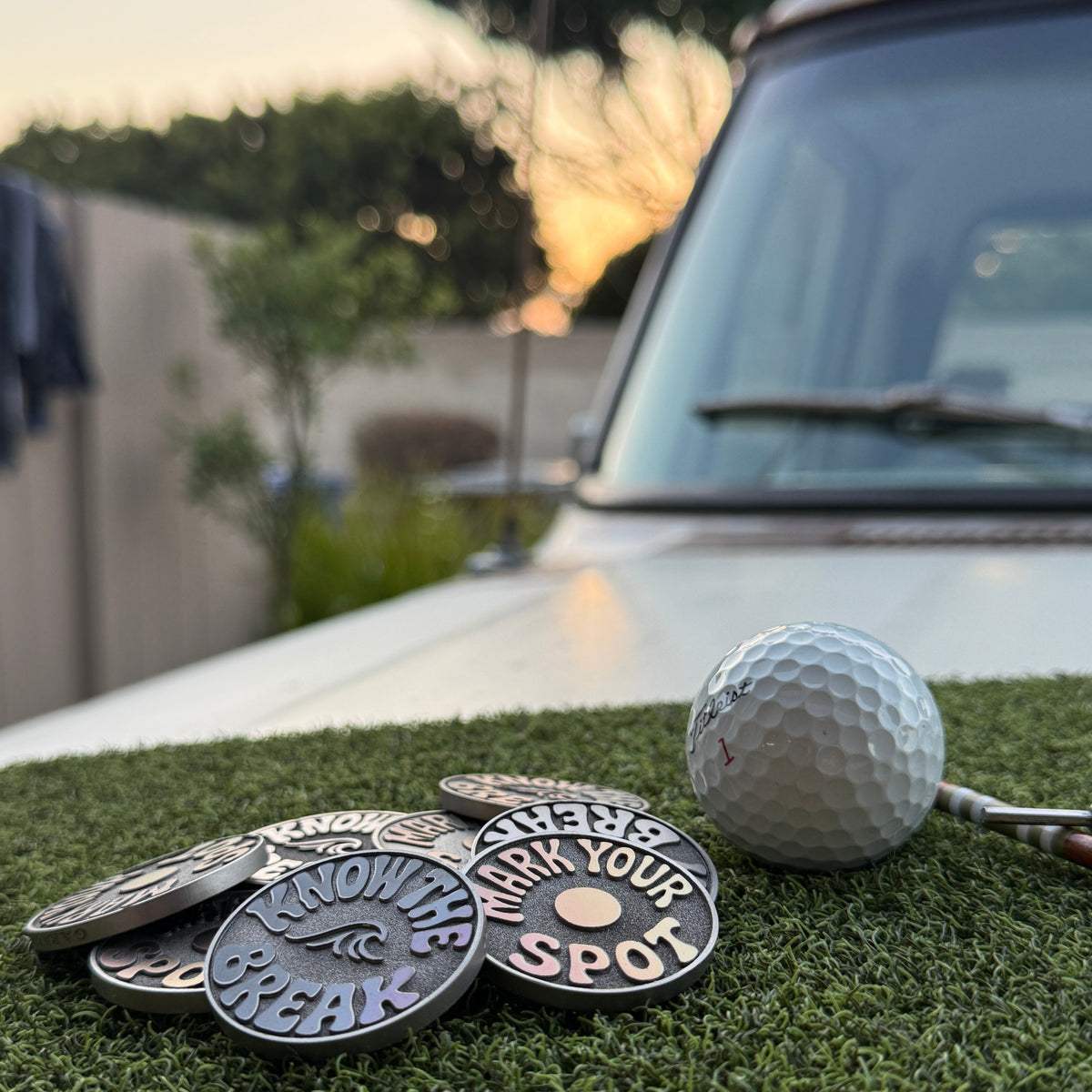 Garage Drop 001 stainless steel golf ball markers by Swell Marks displayed with a golf ball on putting turf at sunset