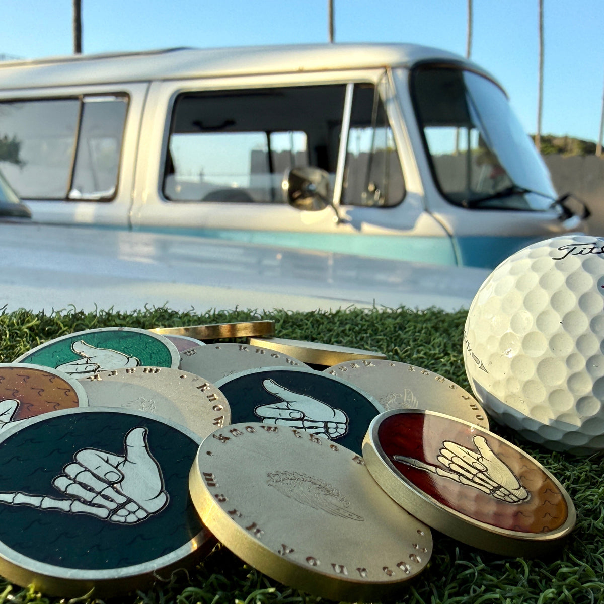 Good Bones brass golf ball markers with colored surfboard resin and skeletal shaka design beside a golf ball and vintage VW Bus.