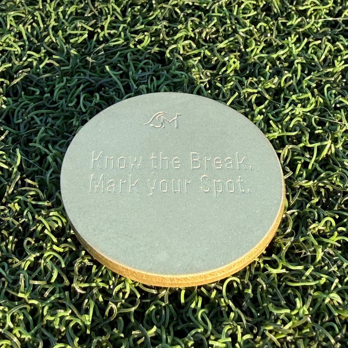 Back of the Lightning brass golf ball marker engraved with the Swell Marks logo and “Know the Break. Mark Your Spot.” on green turf