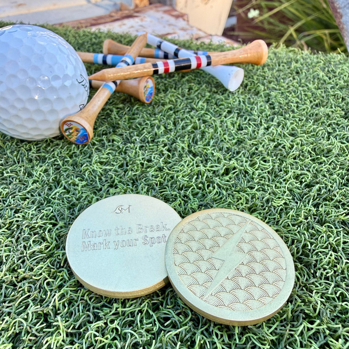 Front and back of the Lightning brass golf ball marker with a raised lightning bolt, Japanese wave pattern, and Swell Marks engraving on green turf
