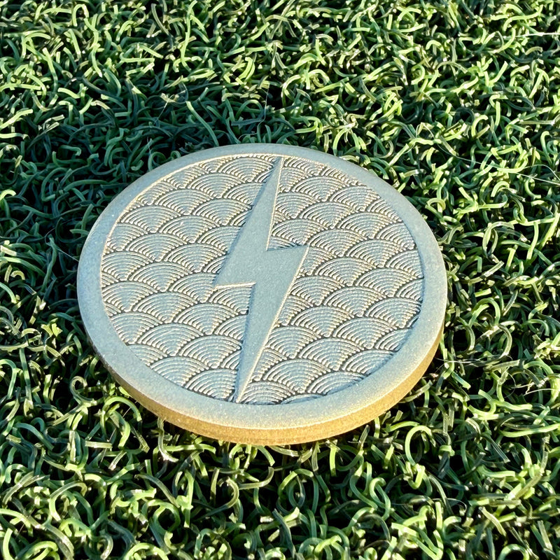 Lightning brass golf ball marker with a raised lightning bolt over an engraved Japanese wave pattern on green turf