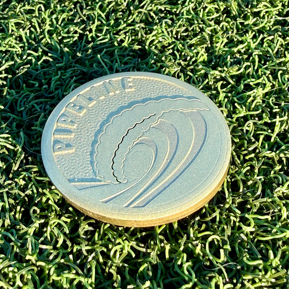 Pipeline brass golf ball marker with a deep engraved breaking wave design photographed on green turf