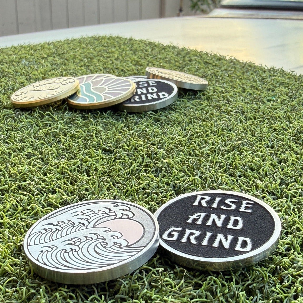 Front and reverse of the Rise & Grind Red Sun stainless steel golf ball marker displayed on green turf with other Swell Marks markers in the background.