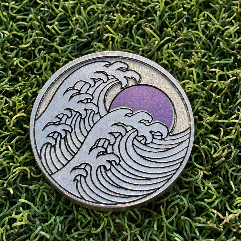 Rise & Grind Red Sun stainless steel golf ball marker with a laser-colored sun and breaking-wave design on green turf.