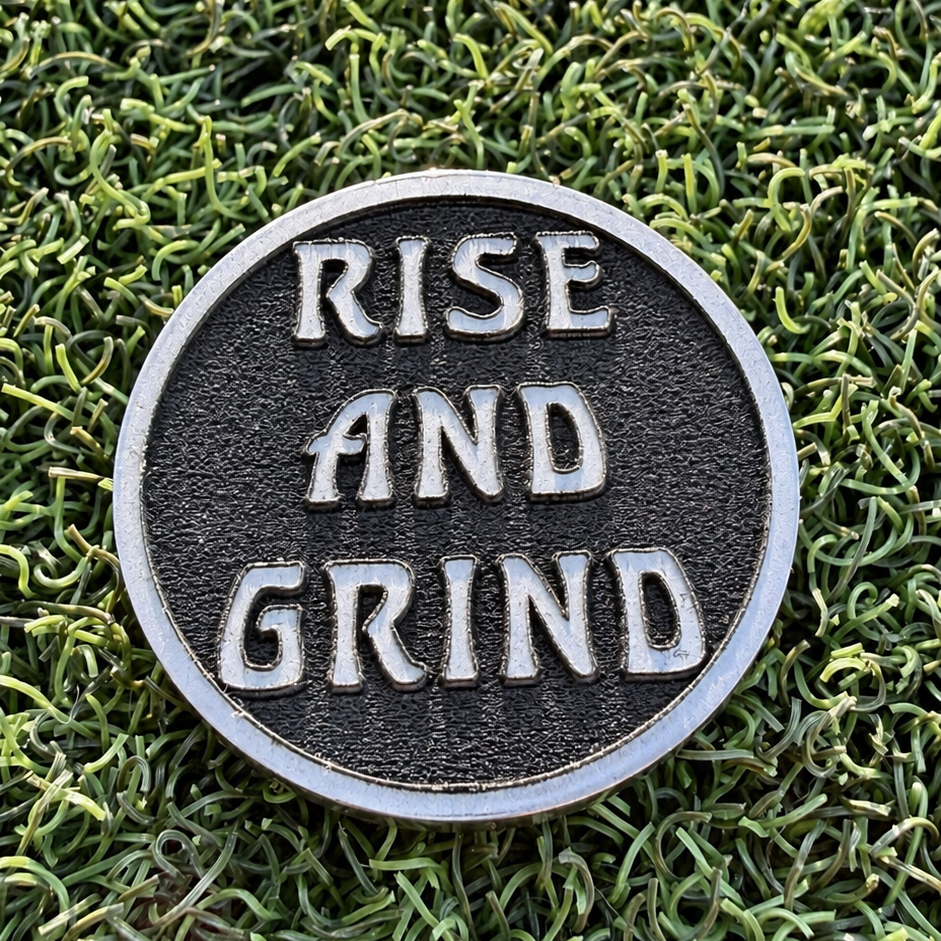 Reverse of the Rise & Grind Red Sun stainless steel golf ball marker with raised lettering on a dark textured background.