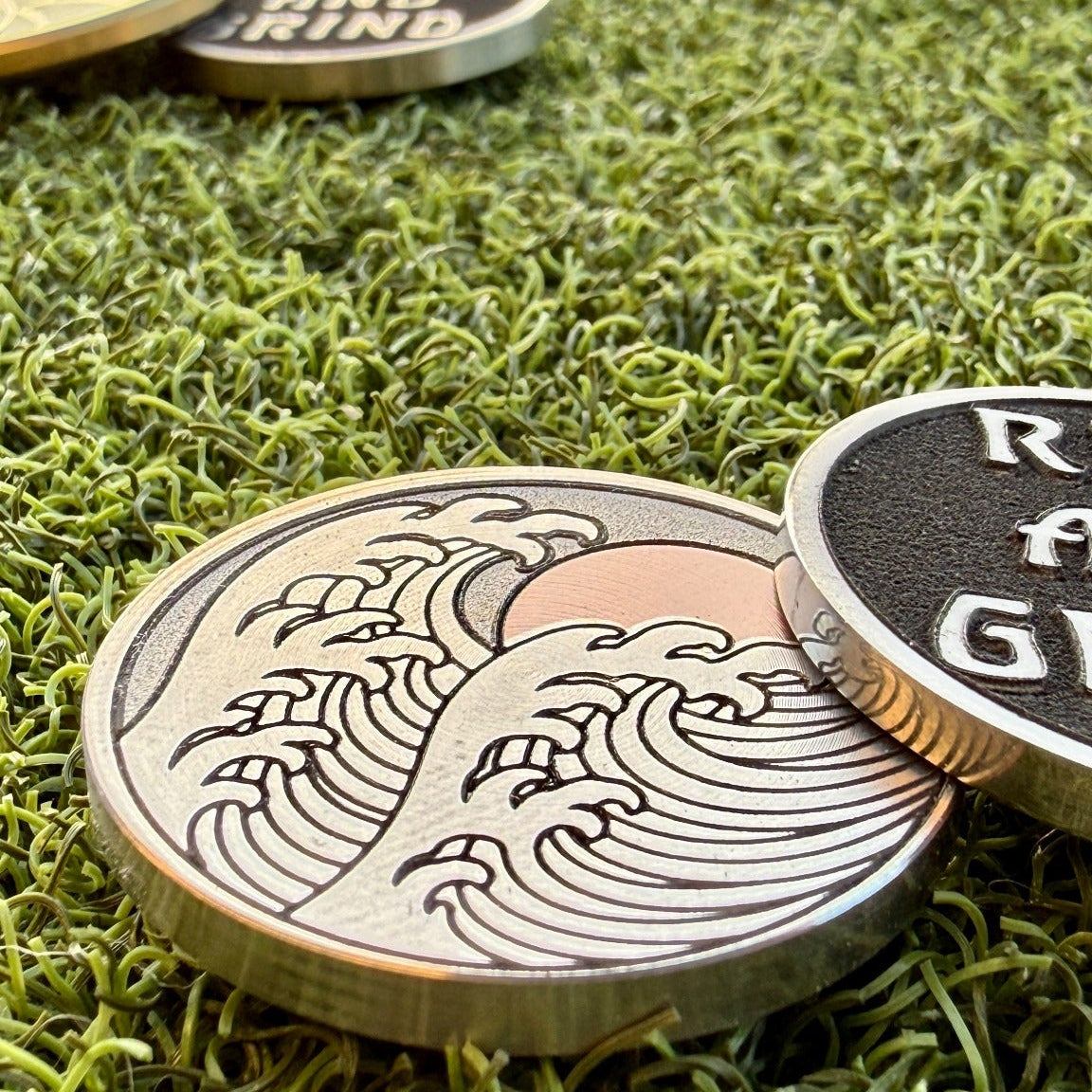 Close-up of the Rise & Grind Red Sun stainless steel golf ball marker with its wave face and raised-letter reverse displayed on green turf.