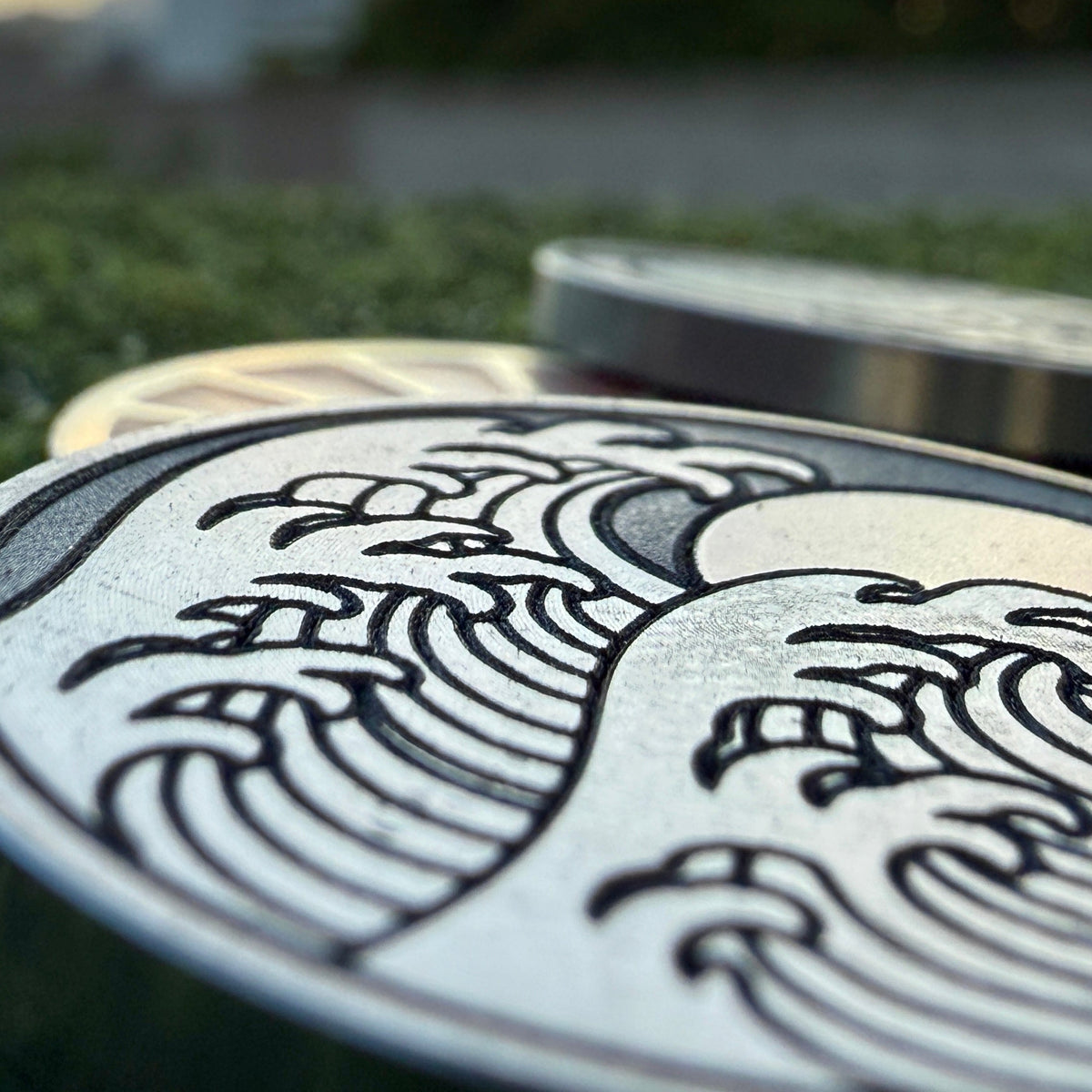 Macro view of the engraved breaking-wave details on the Rise & Grind Red Sun stainless steel golf ball marker.