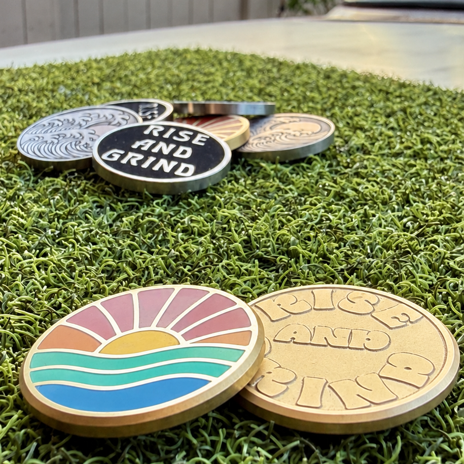 Front and reverse of the Rise & Grind Retro Sunrise brass golf ball marker displayed on green turf.