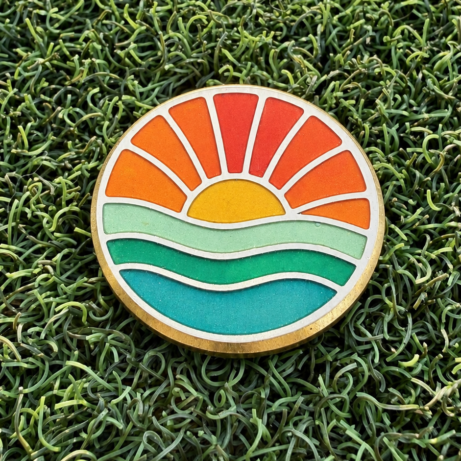 Rise & Grind Retro Sunrise brass golf ball marker with colorful enamel sunrise and ocean design on green turf.
