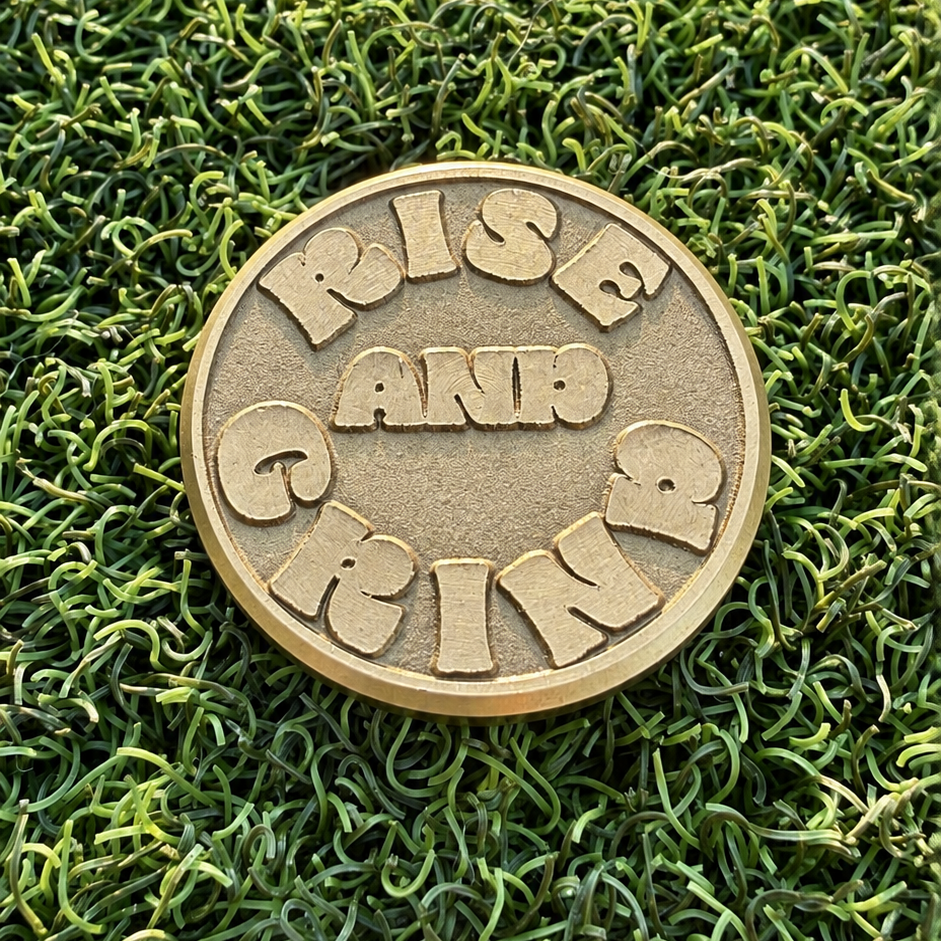 Reverse of the Rise & Grind Retro Sunrise brass golf ball marker with raised lettering on green turf.