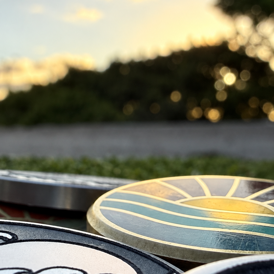 Close-up of the Rise & Grind Retro Sunrise brass golf ball marker with multicolor enamel waves against a warm sunset background.