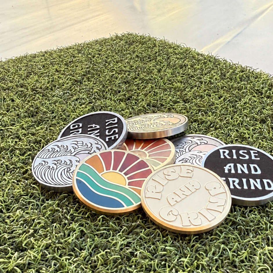 Rise & Grind Retro Sunrise brass golf ball marker displayed with a collection of Swell Marks markers on green turf.