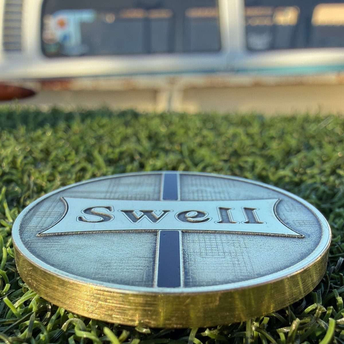 Close-up of the Swell Marks Weathered Glass brass golf ball marker showing the raised Swell badge, resin stringer, and textured finish